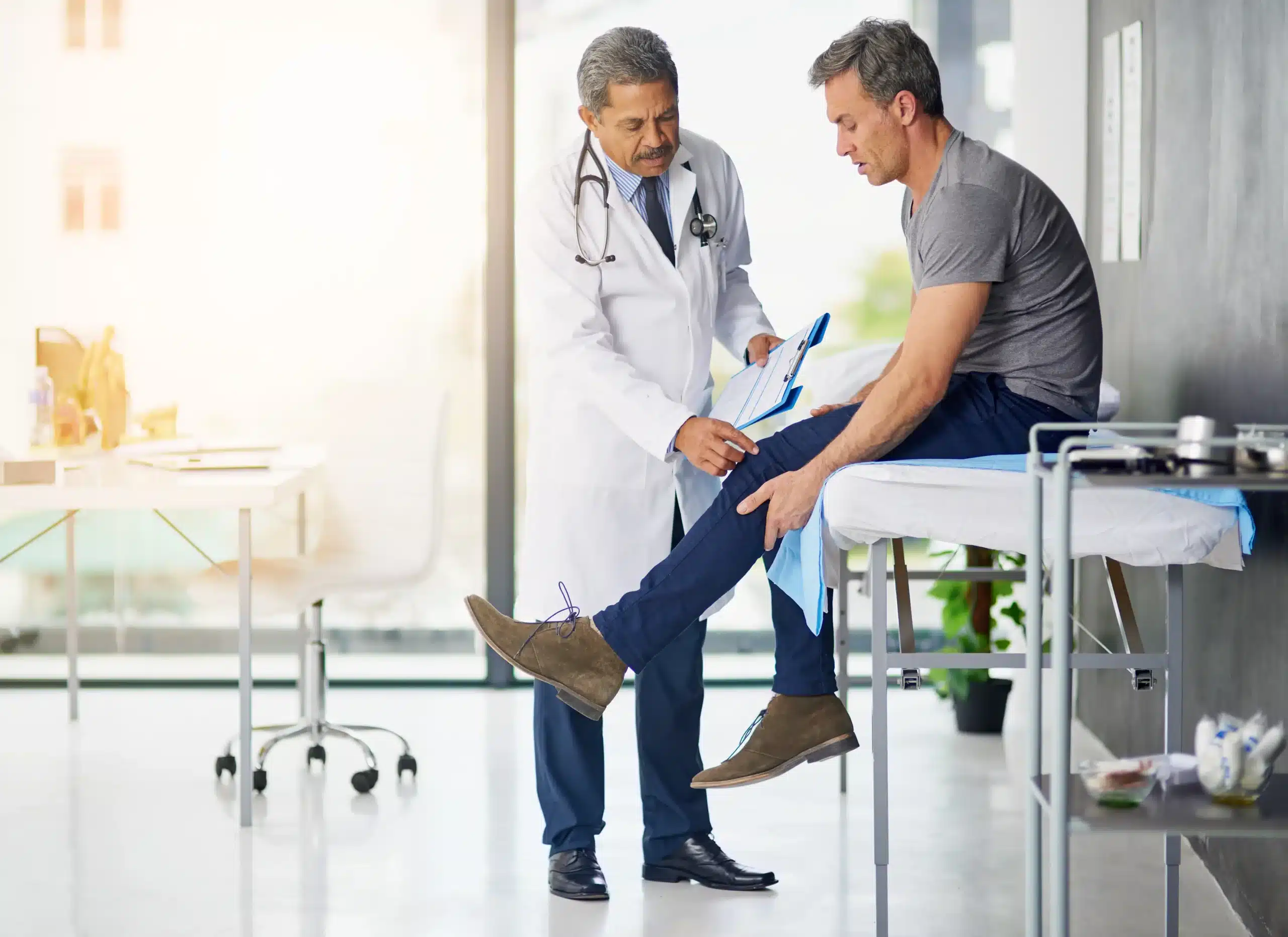 Mature doctor examining a concerned patient’s knee during an independent medical exam to assess work-related injury and recovery progress.