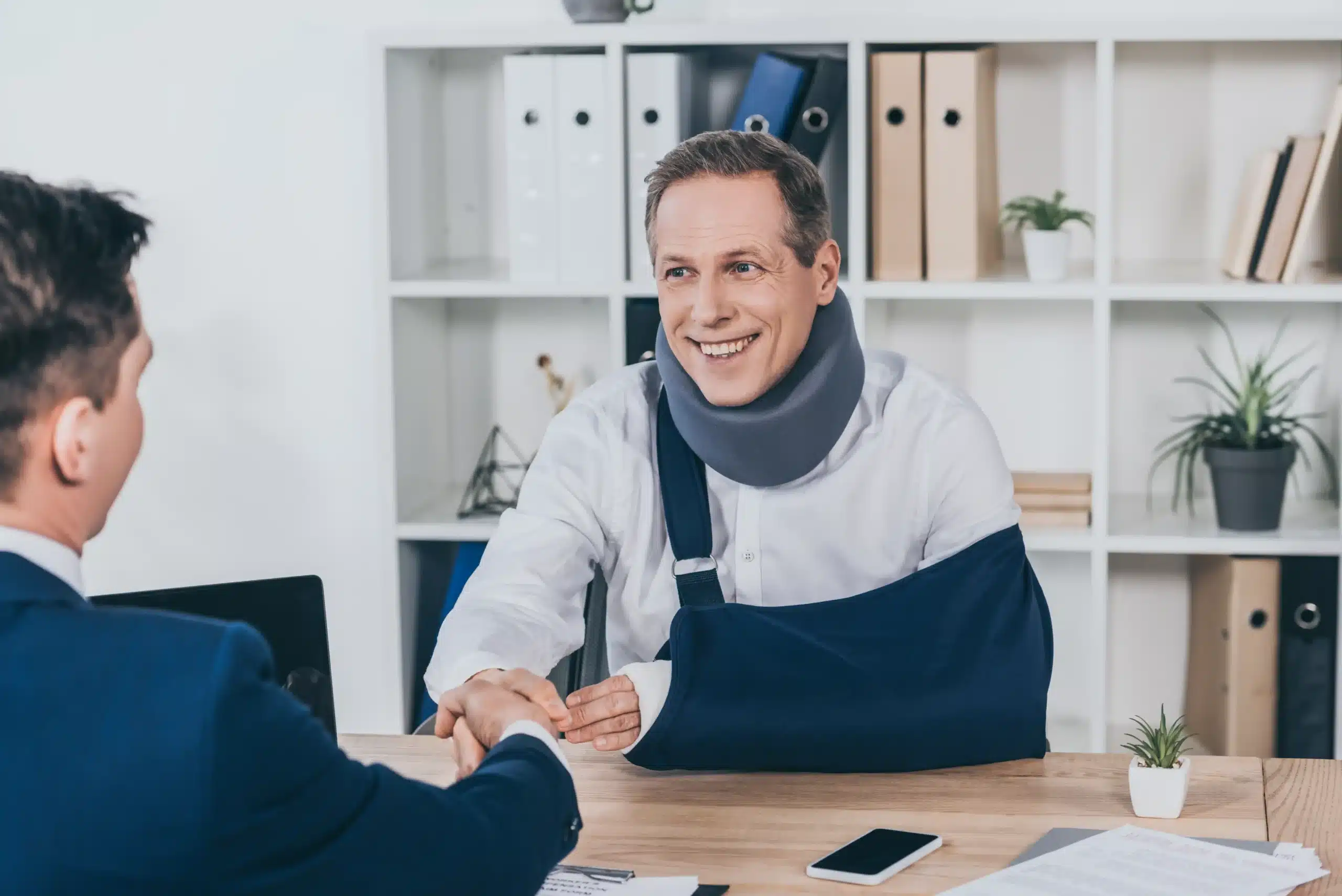 Injured worker wearing neck brace and arm cast shaking hands with lawyer in New York office discussing workers’ compensation benefits.